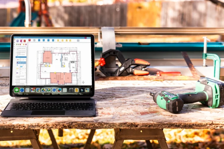 Laptop displaying digital house plans on a construction workbench next to a cordless drill, with building tools and materials in the background.