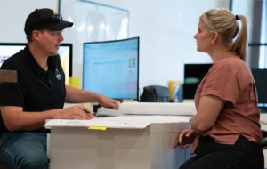 Two people talking next to a desktop computer.