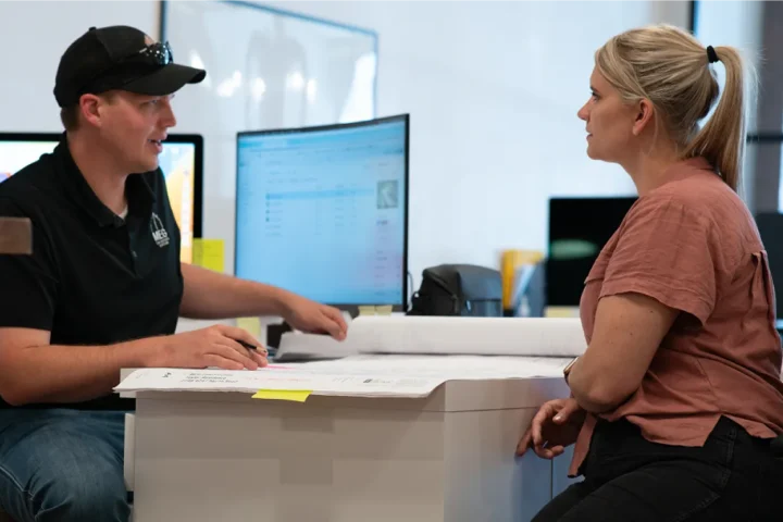 Two people talking next to a desktop computer.