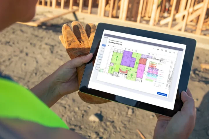Construction worker holding a tablet displaying a color-coded floor plan on the Square Takeoff app at a building site under construction.