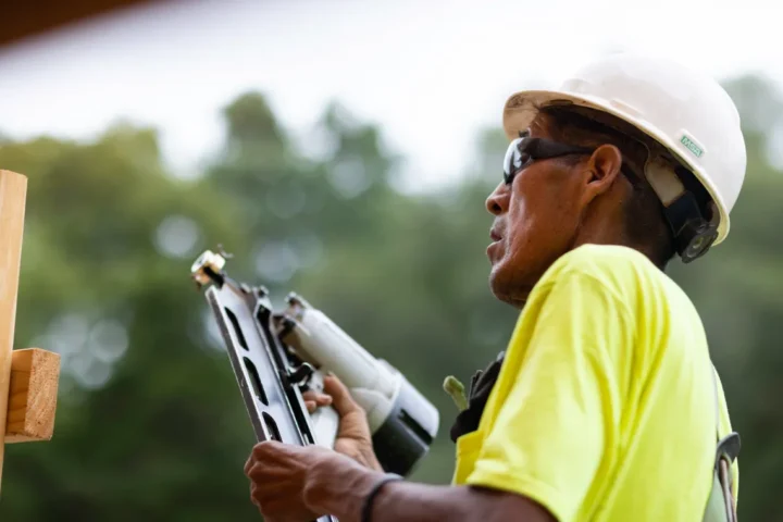 Construction worker wearing a hard hat and safety glasses using a nail gun to fasten framing on a residential build site.